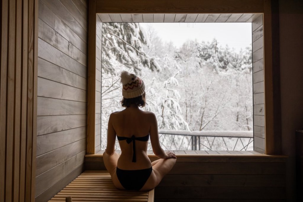 woman in a beanie in sauna looking at the window with snowy trees